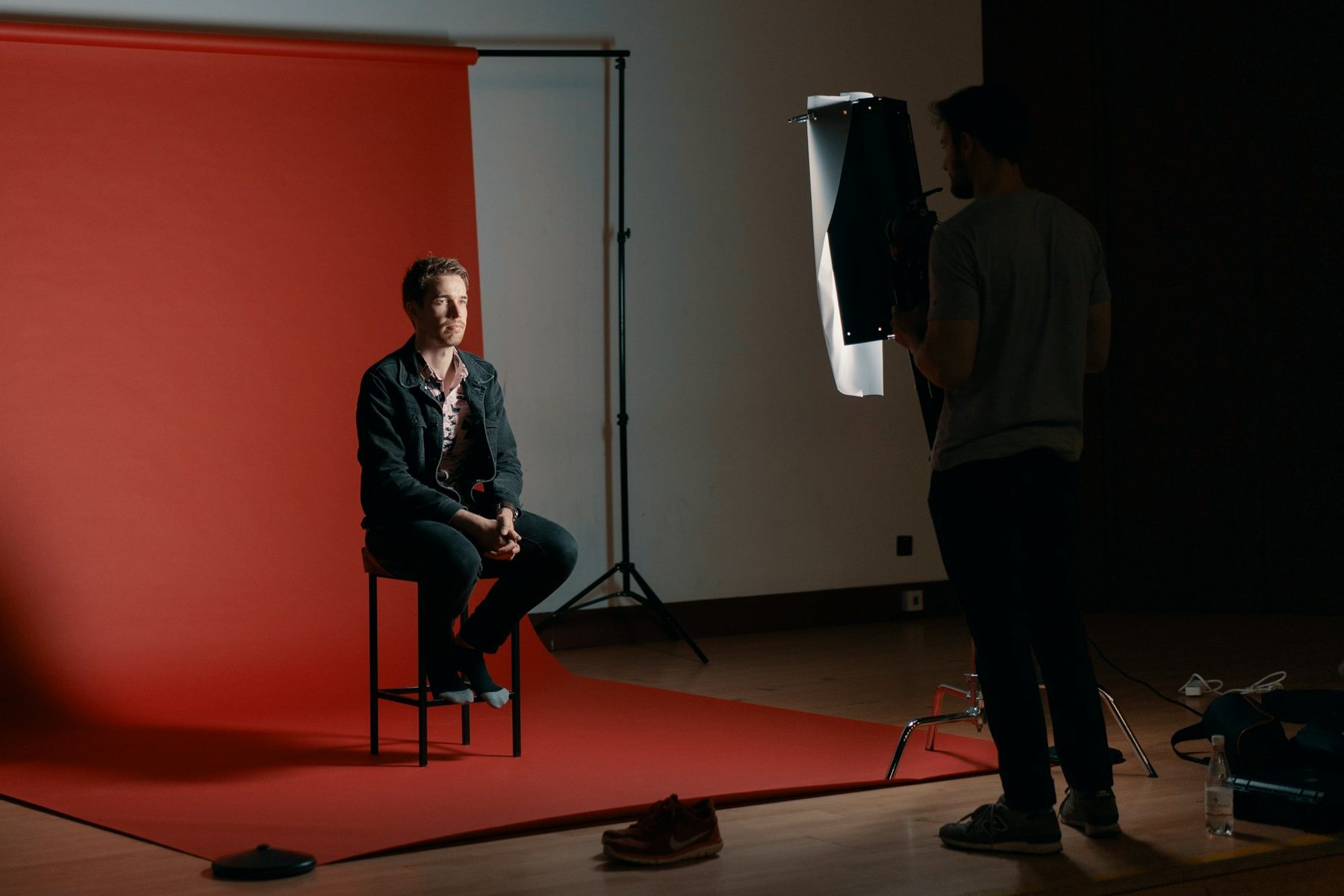 Man seated on a stool against a red studio backdrop while a crew member adjusts a light