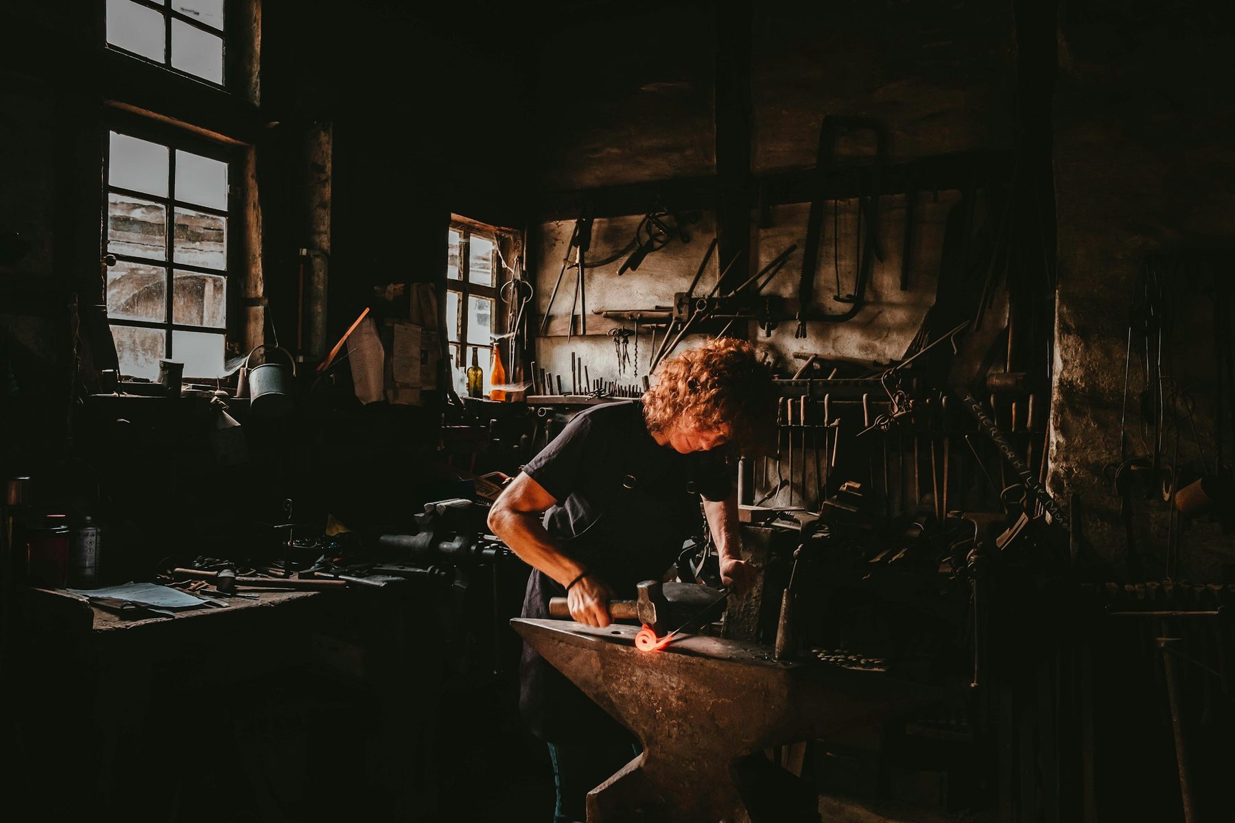 Blacksmith working at an anvil in a dark forge lit by warm window light