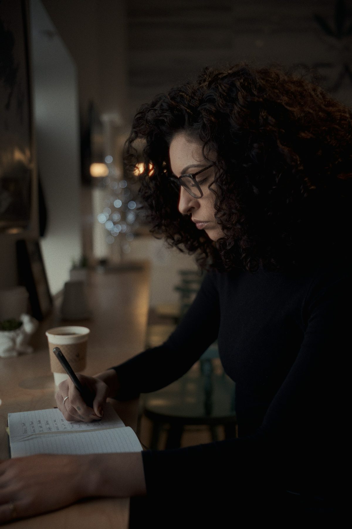 Woman writing notes in a notebook at a dark cafe counter beside a coffee cup