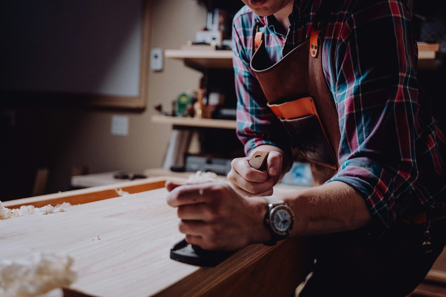 Craftsman in a leather apron planing wood at a workbench in a dimly lit workshop