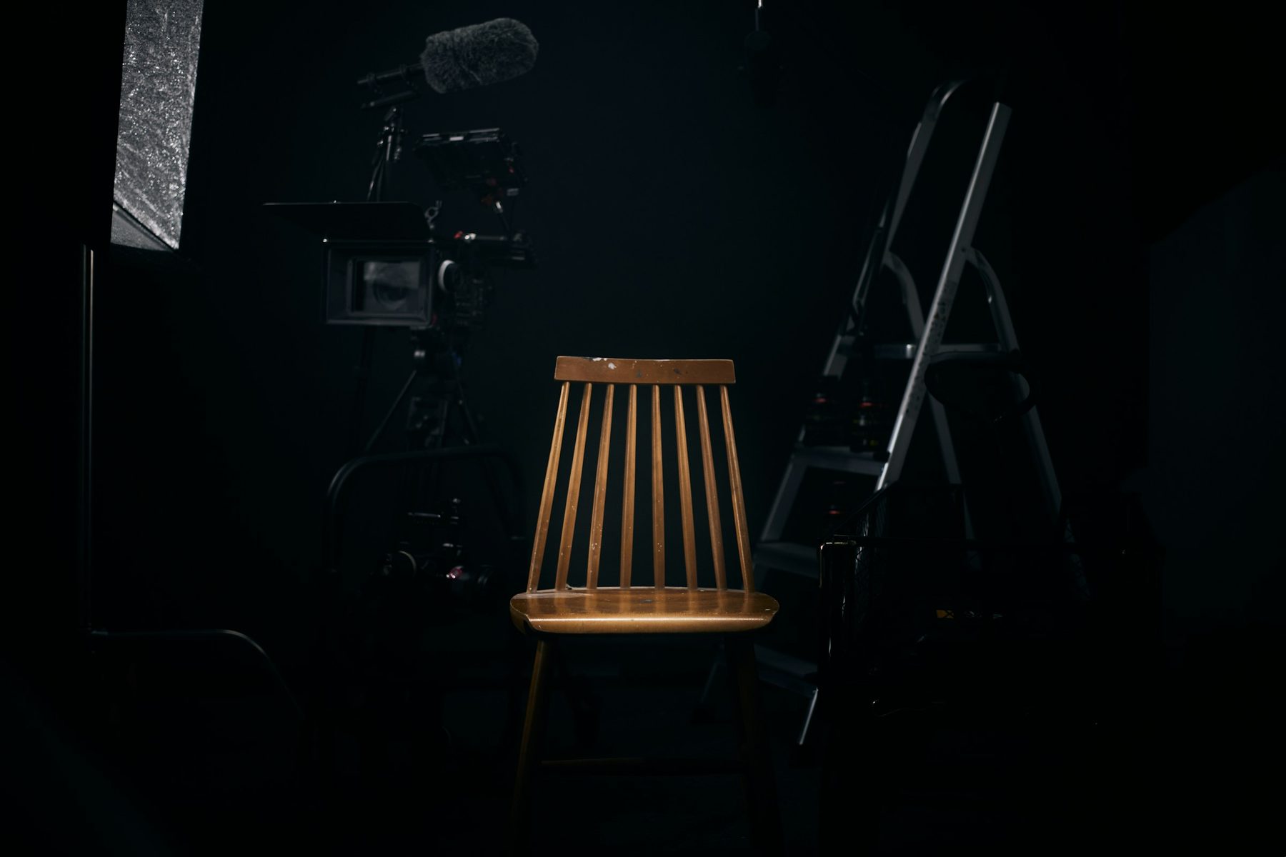 Empty wooden chair lit on a dark studio set with a cinema camera and boom microphone waiting behind it