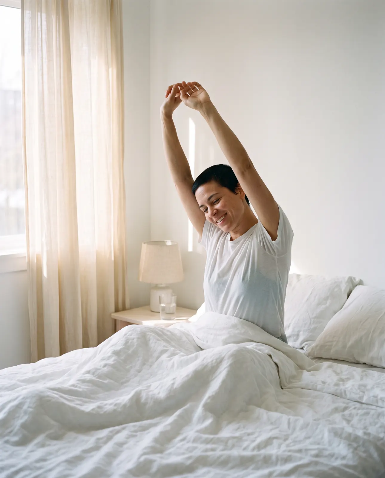A person sits up in bed in morning light, stretching and smiling.