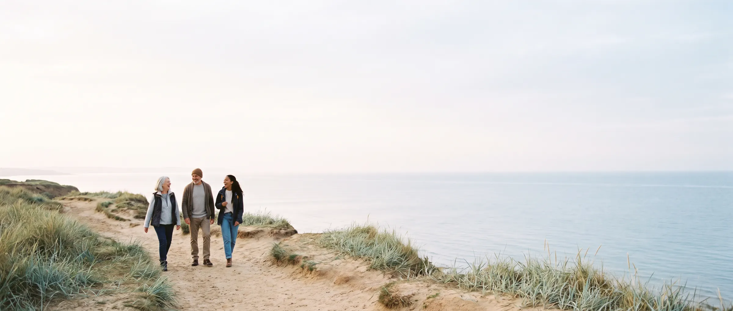 Three friends of different ages walk and laugh along a coastal path above a calm sea.