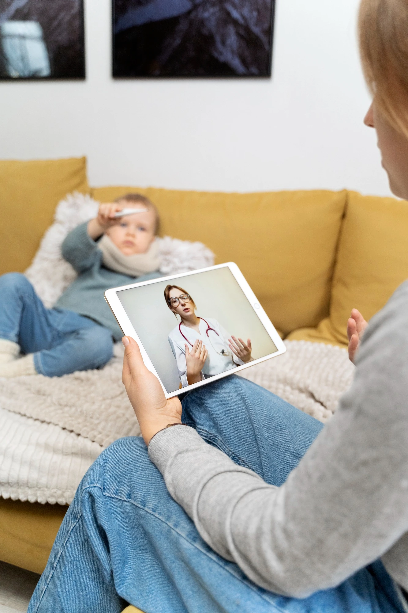 Parent on a tablet video call with a doctor while a sick child checks a thermometer on the sofa