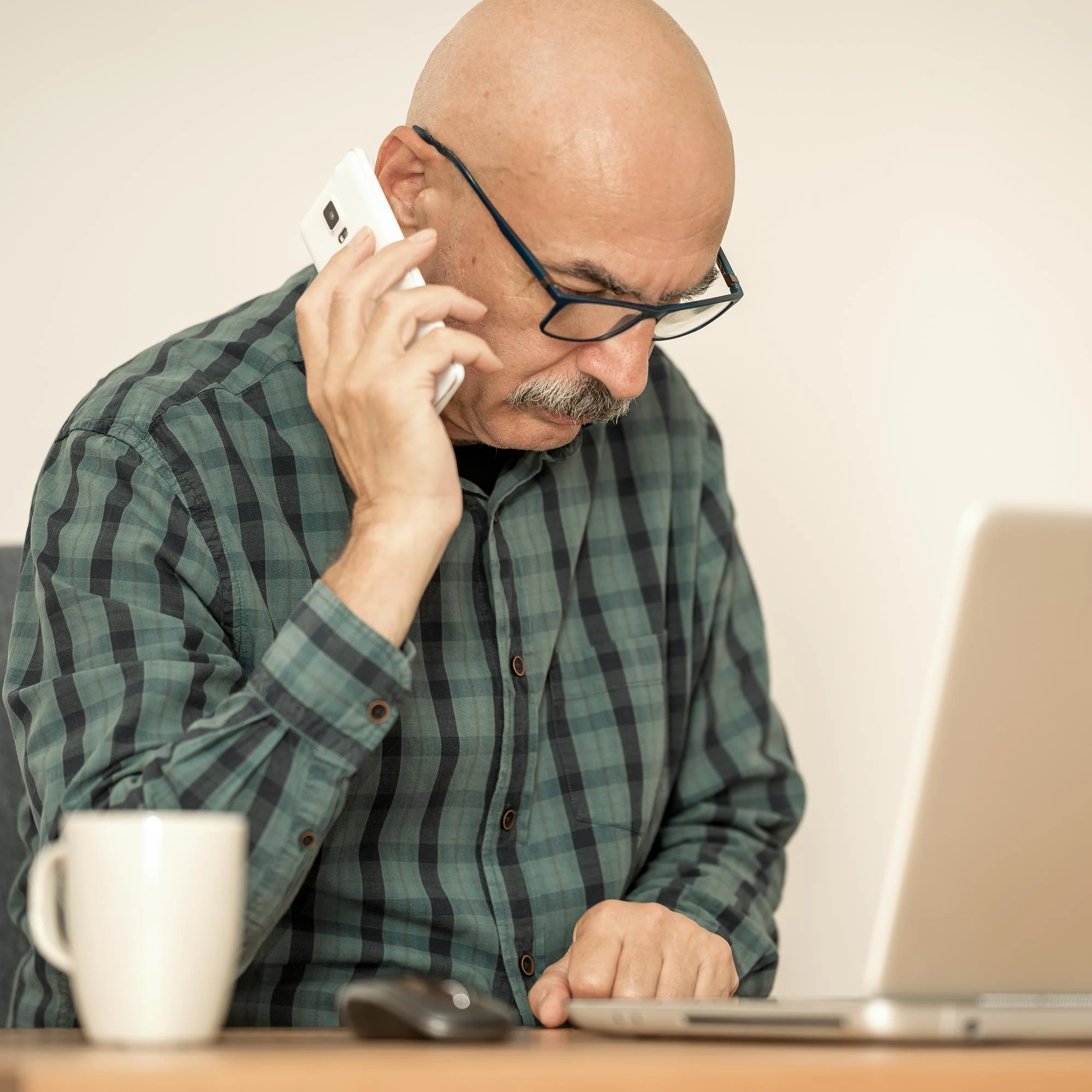 Man with glasses on a phone call while working at a laptop with a coffee mug
