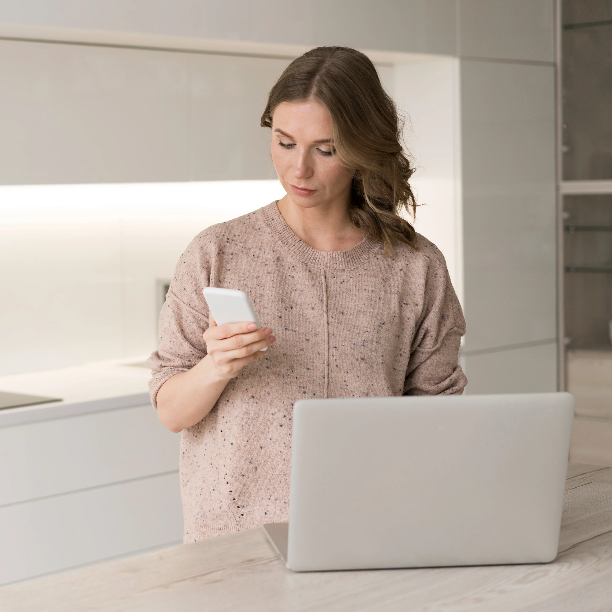 Woman standing at a laptop checking her phone in a bright kitchen