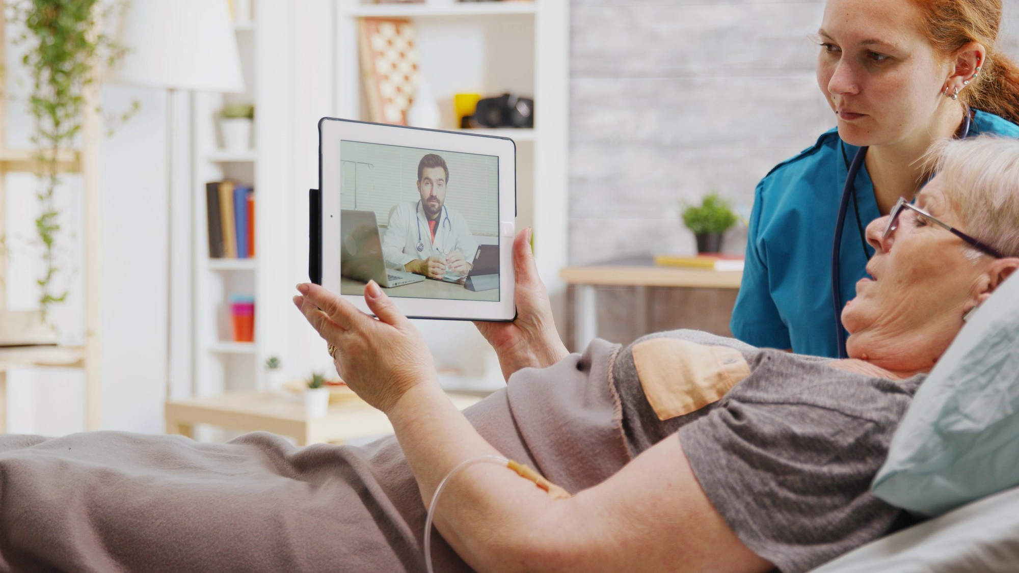 Elderly woman in bed using a tablet to video call a doctor while a nurse assists