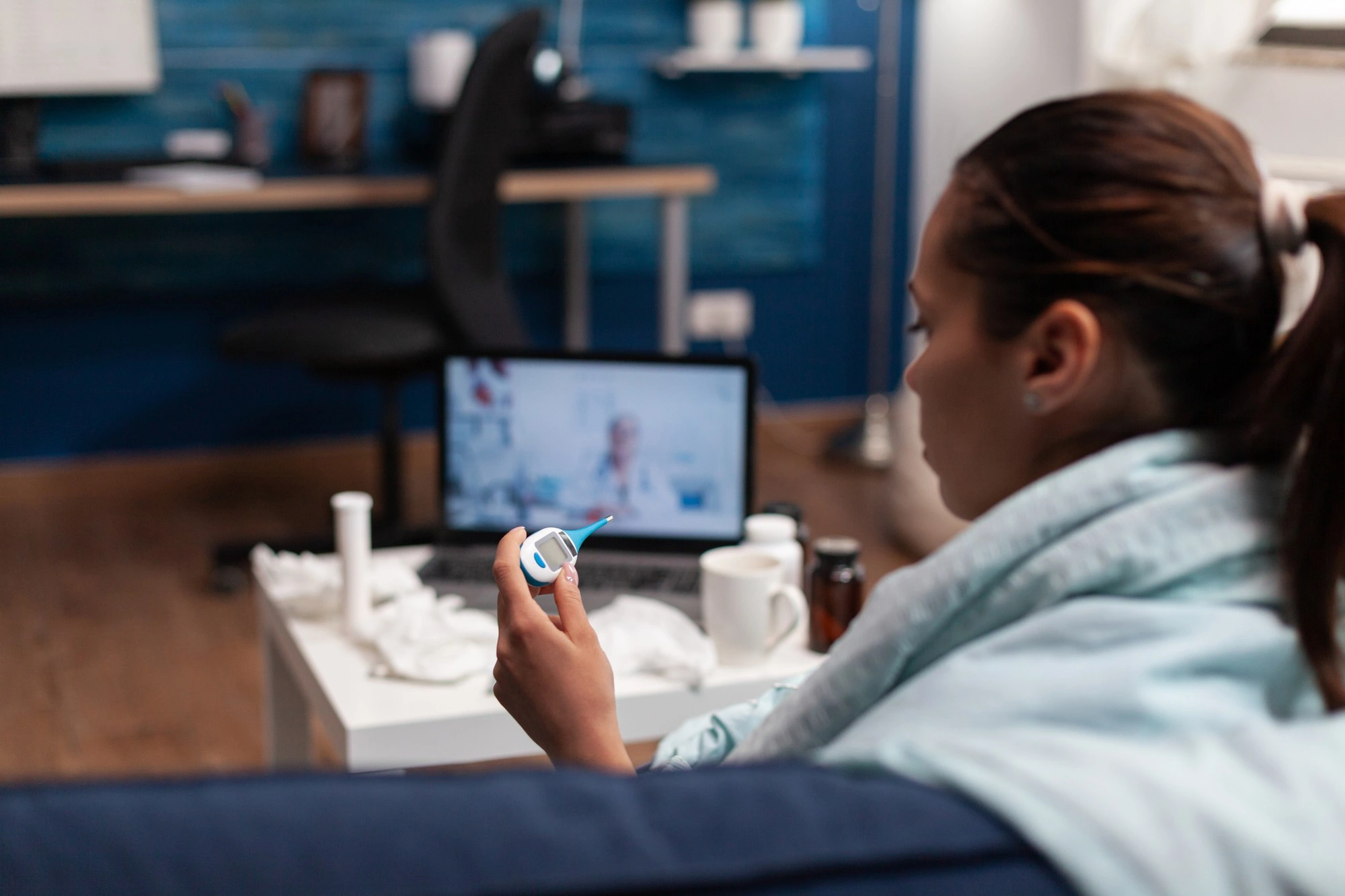 Woman wrapped in a blanket checking her temperature while on a video call with a doctor