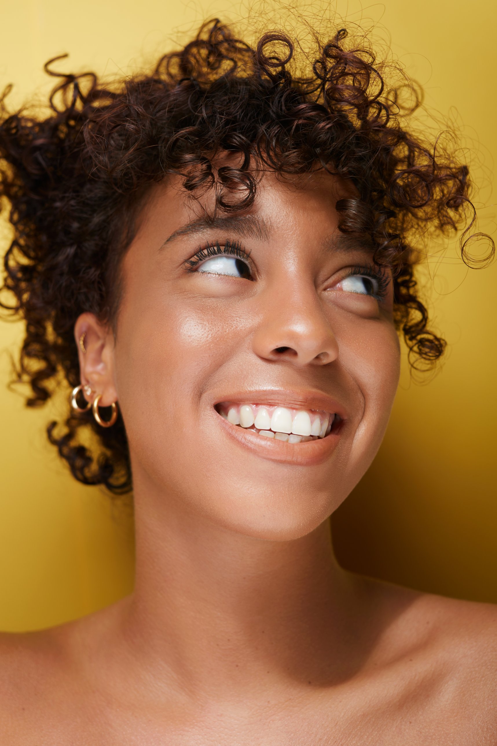 Portrait beauté d'une femme au sourire éclatant, cheveux bouclés, peau retouchée sur fond jaune moutarde