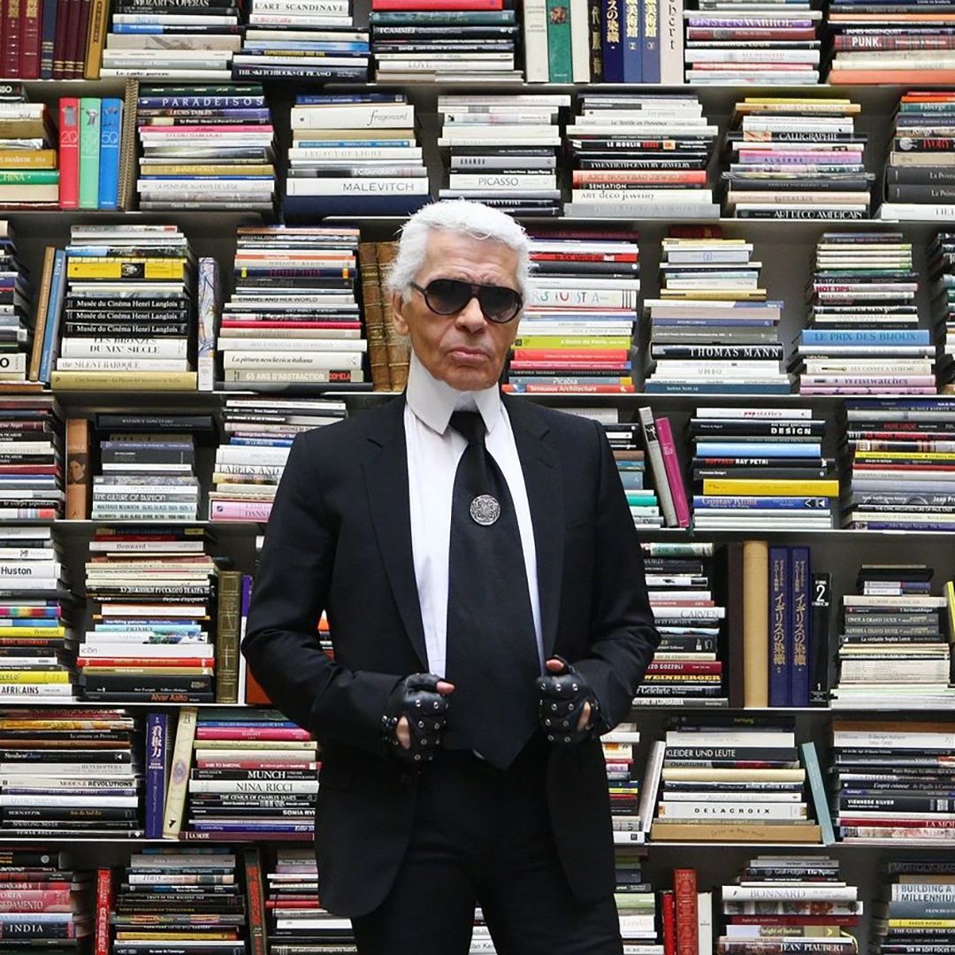 Karl Lagerfeld en costume noir, lunettes sombres et gants, devant une bibliothèque remplie de livres d'art