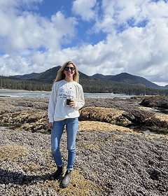 Woman in a white sweatshirt and jeans holding a coffee on a rocky shoreline with forested mountains behind her
