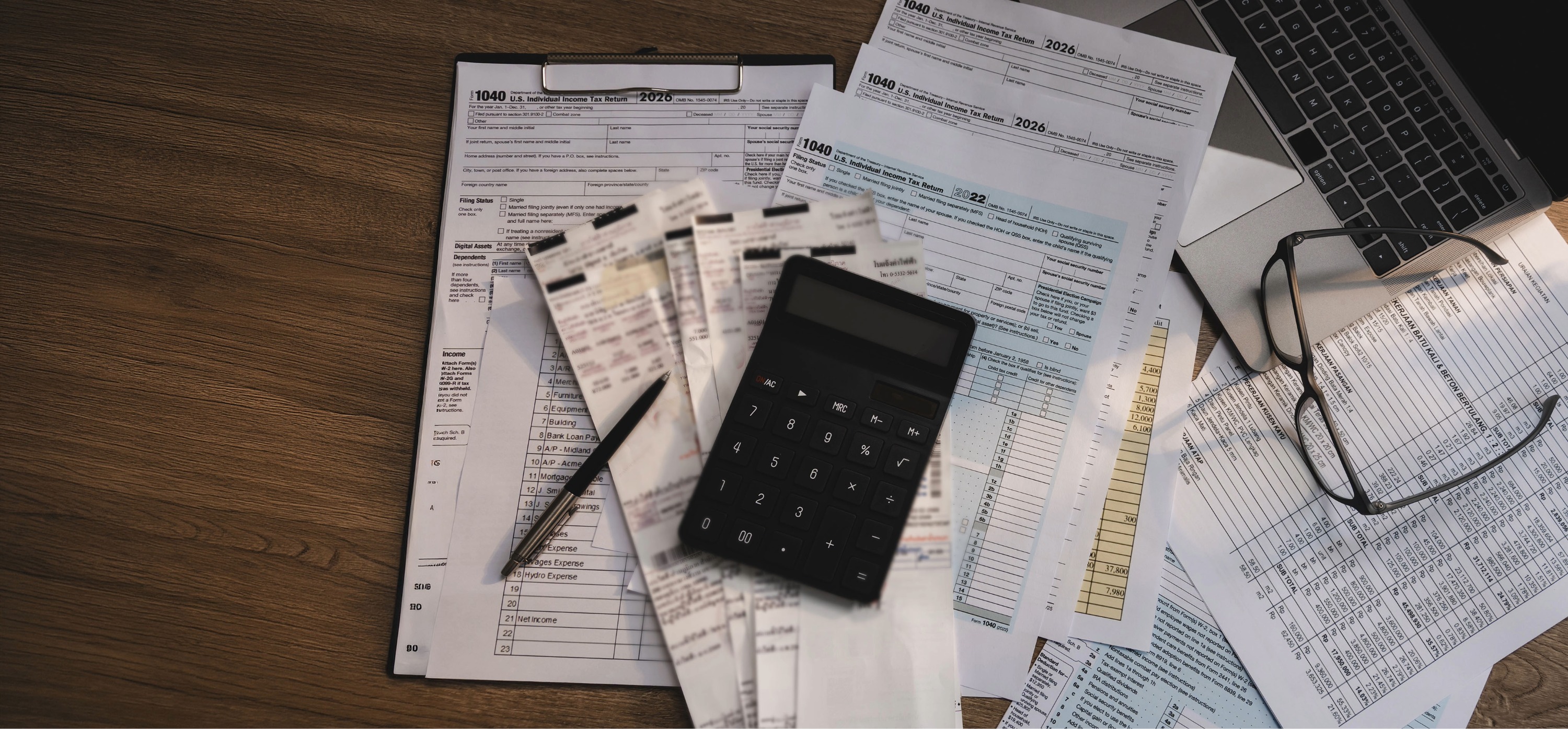 Calculator, pen, use tax forms and receipts scattered on a wooden desk next to a laptop and eyeglasses.