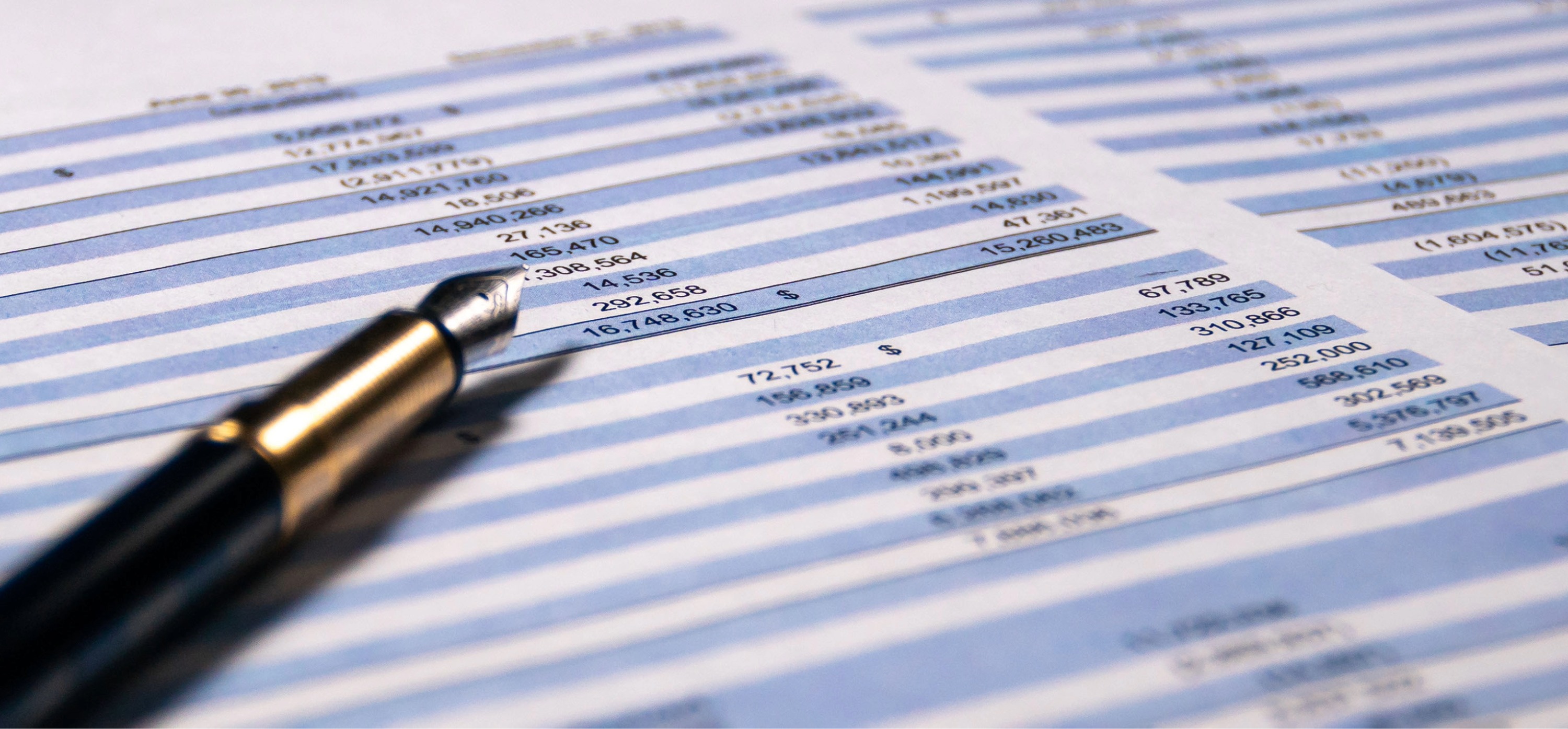 Close-up of a fountain pen resting on printed financial statements with rows of numbers and dollar signs on blue and white striped paper.