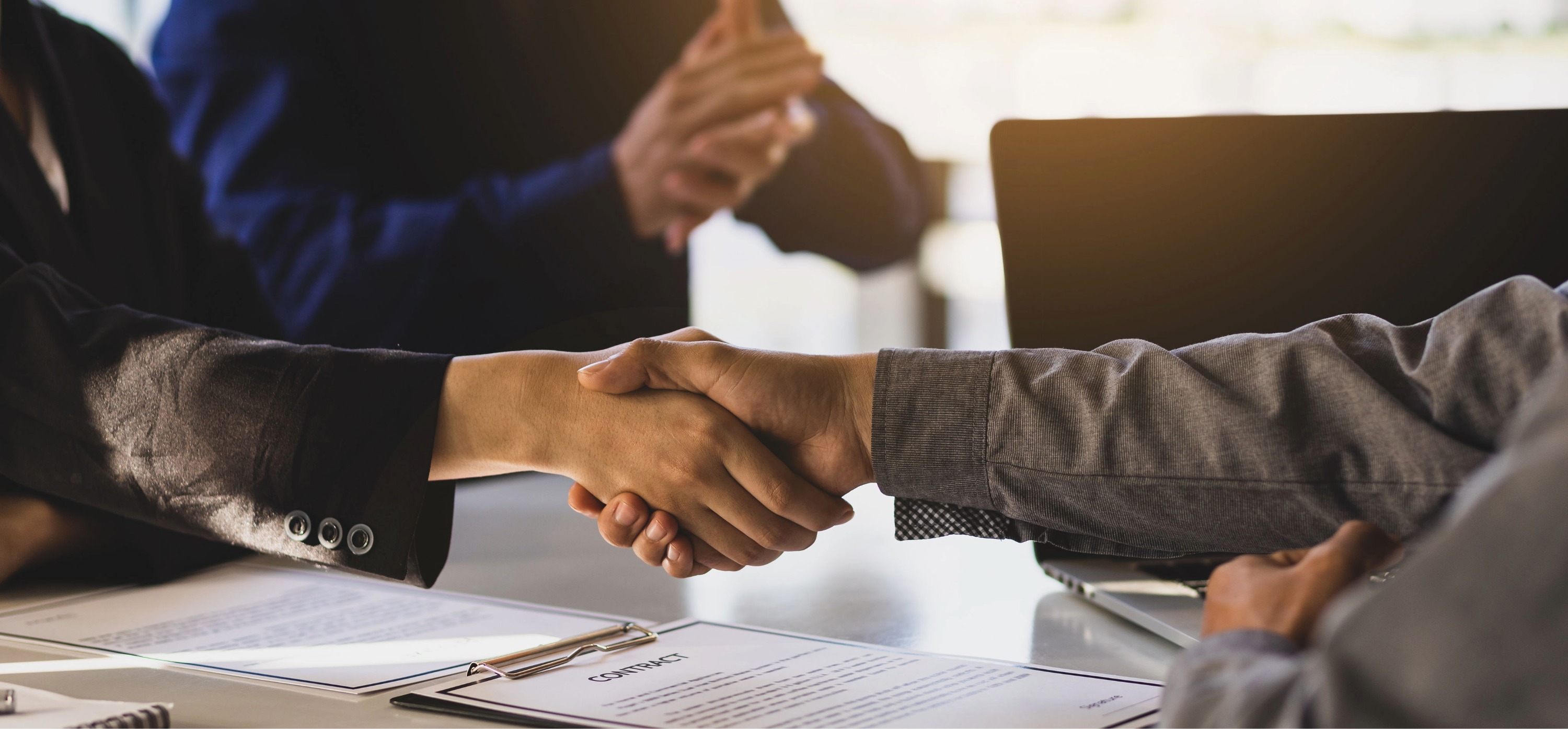 Two people shaking hands over a desk with contract documents, symbolizing a business agreement.