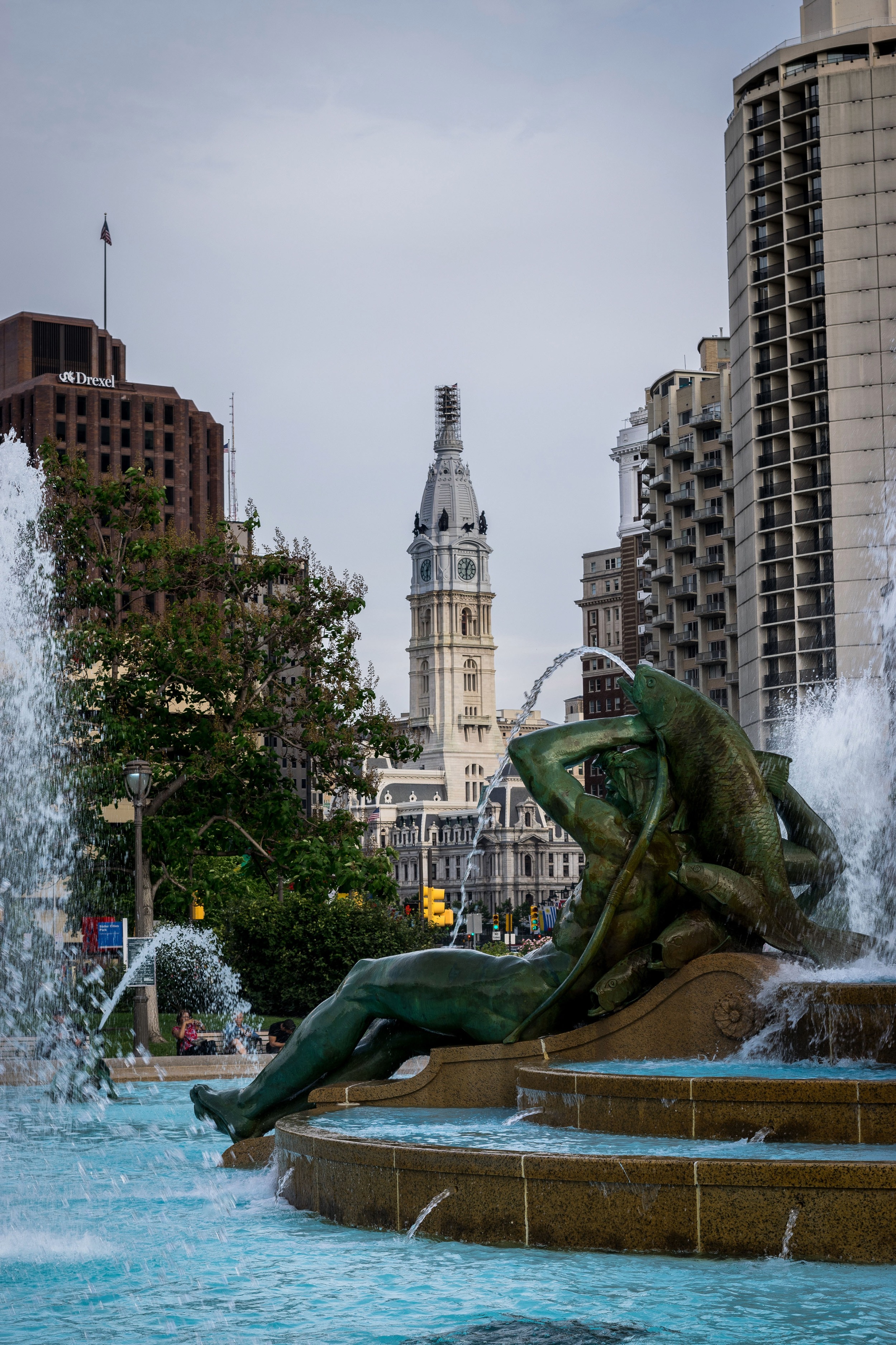 Fountain with sculpture of a reclining figure holding fish that spout water, with Philadelphia City Hall and surrounding buildings in the background.