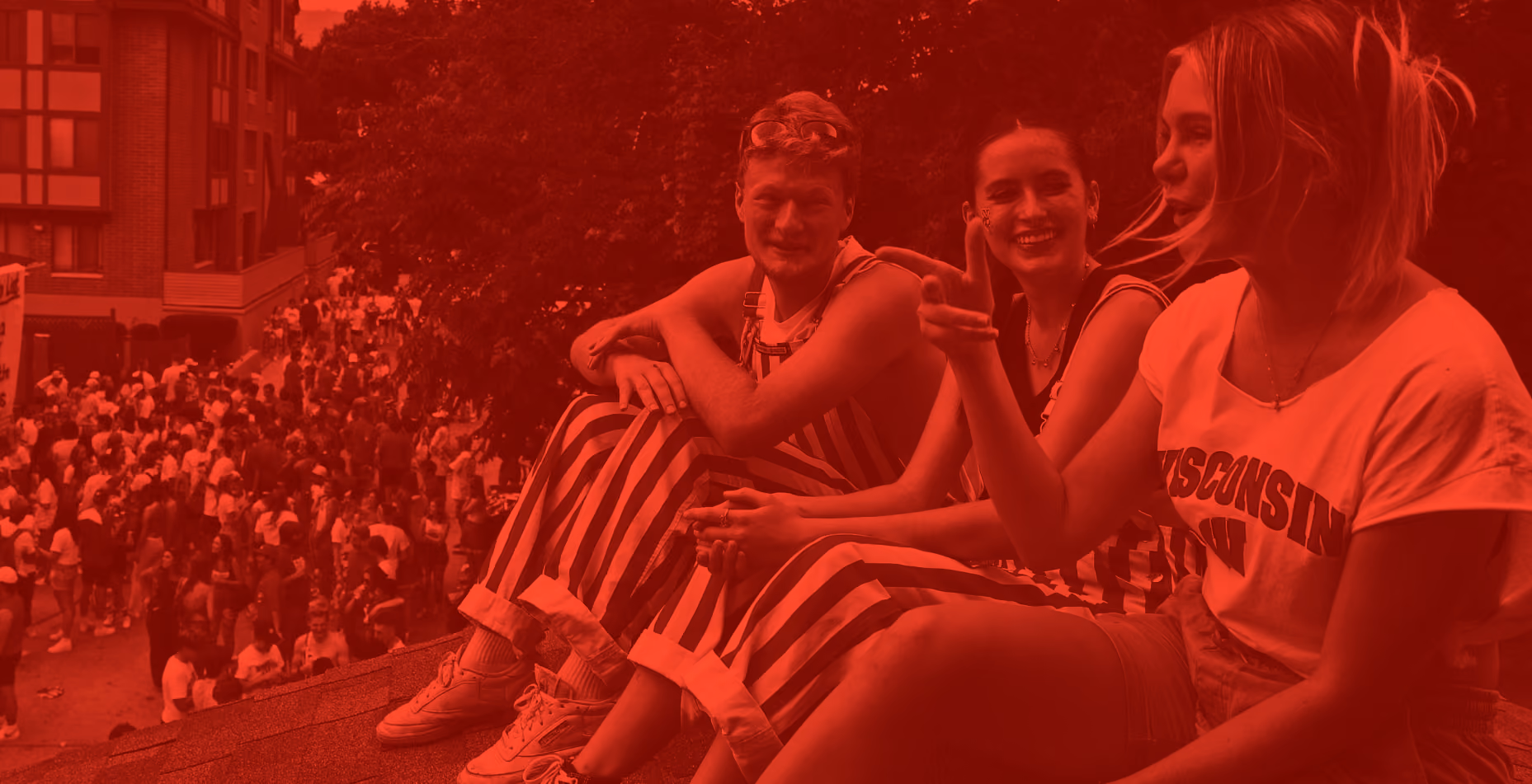 Three students in Wisconsin gear sitting above a game-day crowd in Madison, in a red duotone treatment