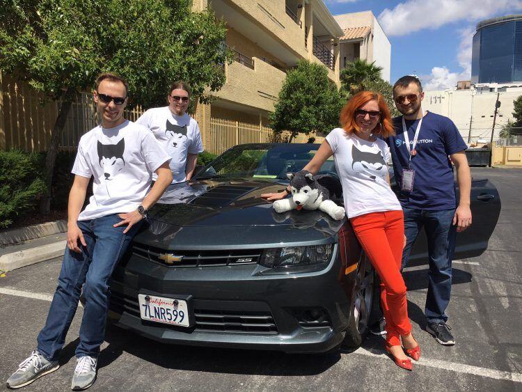 Snowdog team members in husky T-shirts pose around a black Camaro with a plush snow dog on its hood in Las Vegas