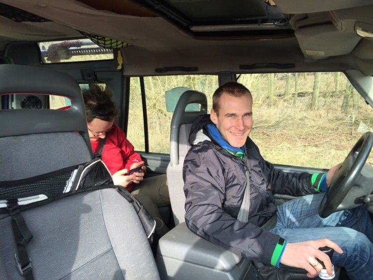Smiling driver at the wheel of a Land Rover, a passenger in a red jacket checking her phone behind him