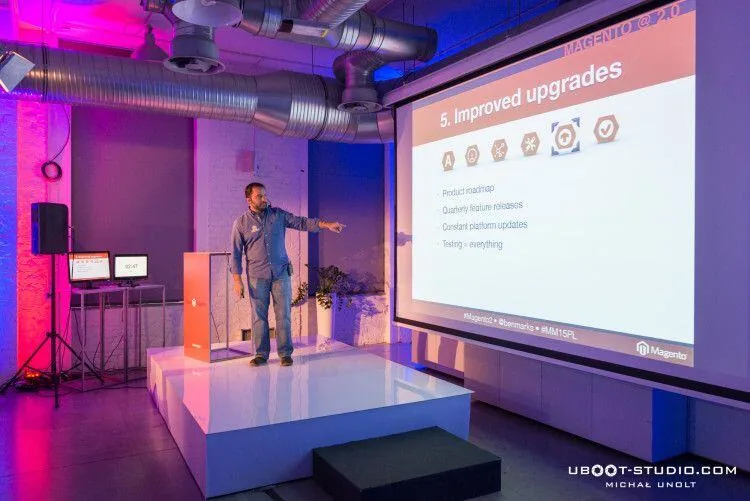 A speaker on stage at Meet Magento Poland points to a Magento 2.0 slide titled "5. Improved upgrades"
