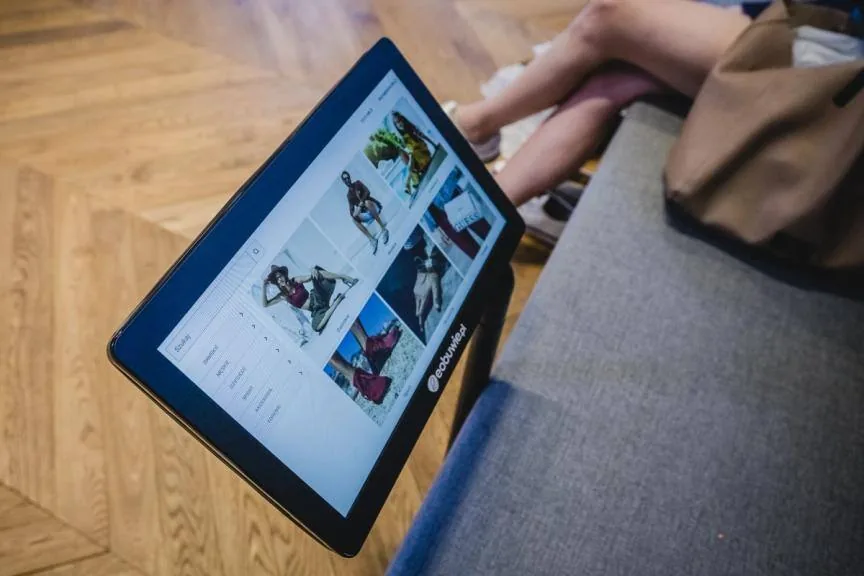 Tablet on a stand beside a bench in the eobuwie.pl store, showing a grid of shoe photos from the store app