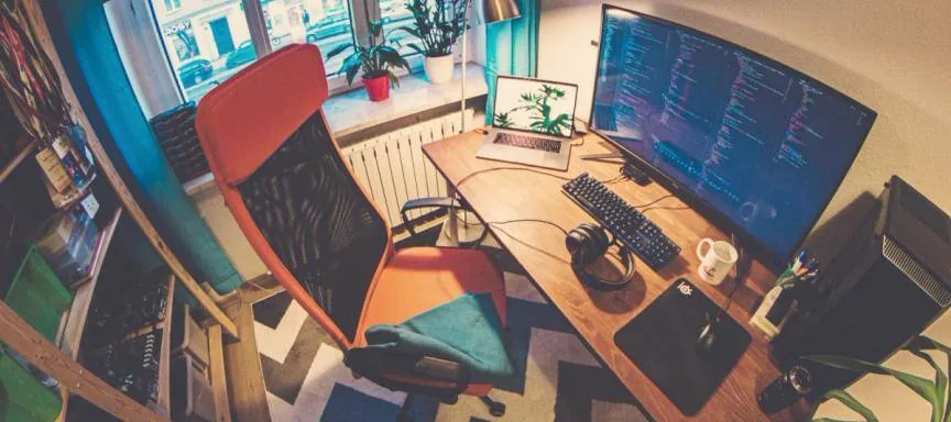 Wide-angle view of Igloczek's home office: orange chair, curved monitor showing code, laptop and plants by the window