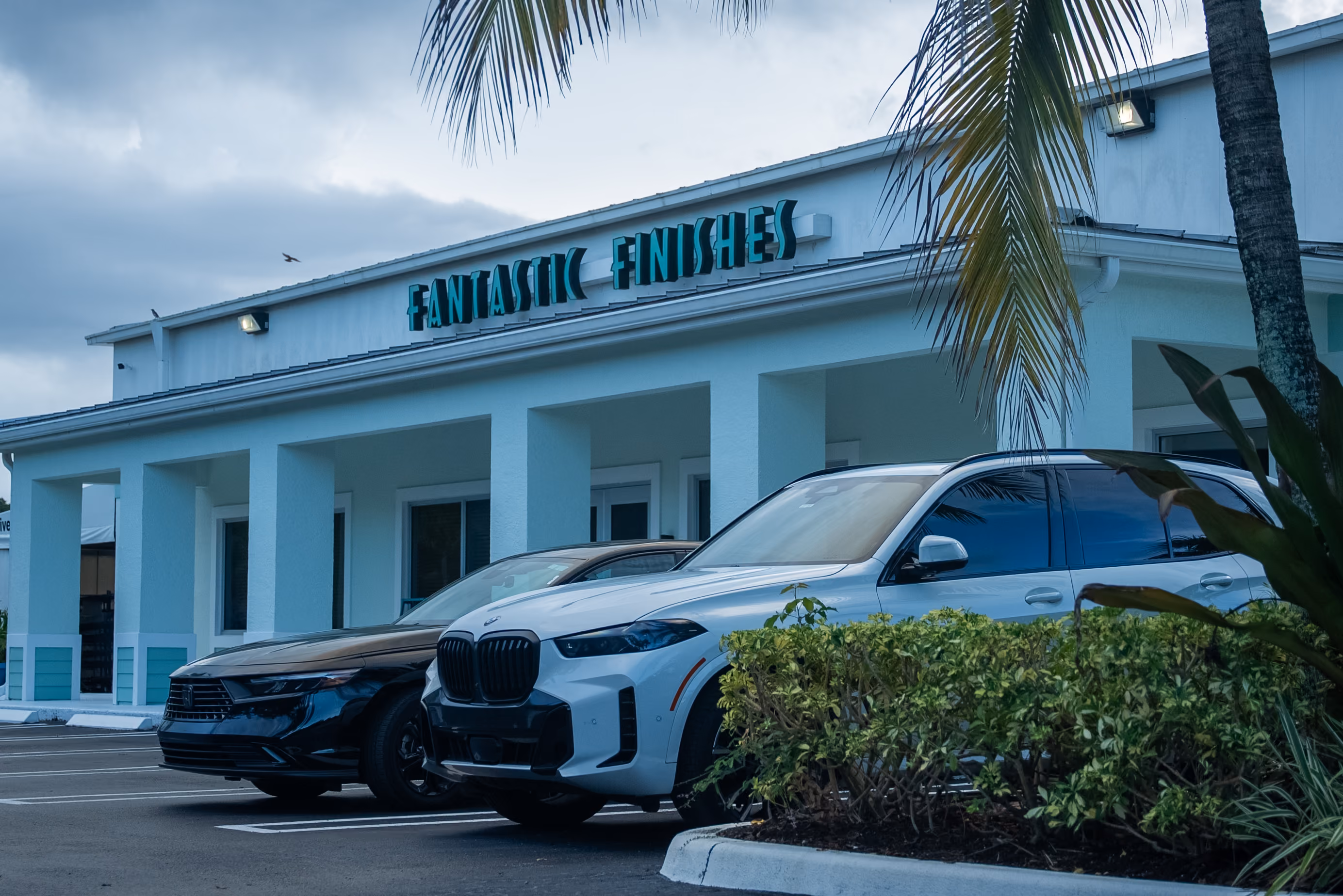 Two parked cars, a black sedan and a white SUV, in front of a light blue building with the sign 'Fantastic Finishes' above the entrance, with palm trees partially visible.