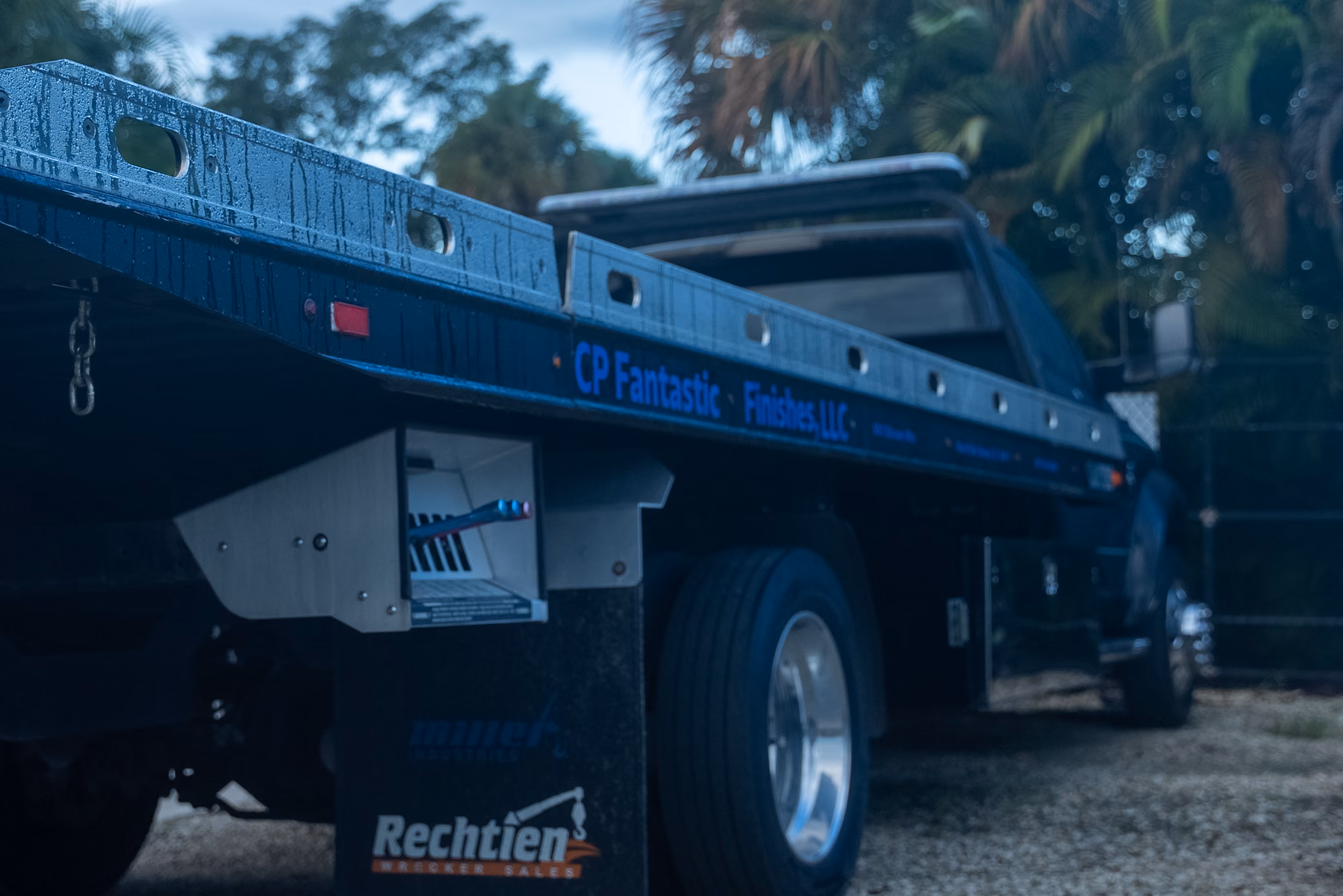 Close-up side view of a blue flatbed tow truck parked on a gravel surface with trees in the background.