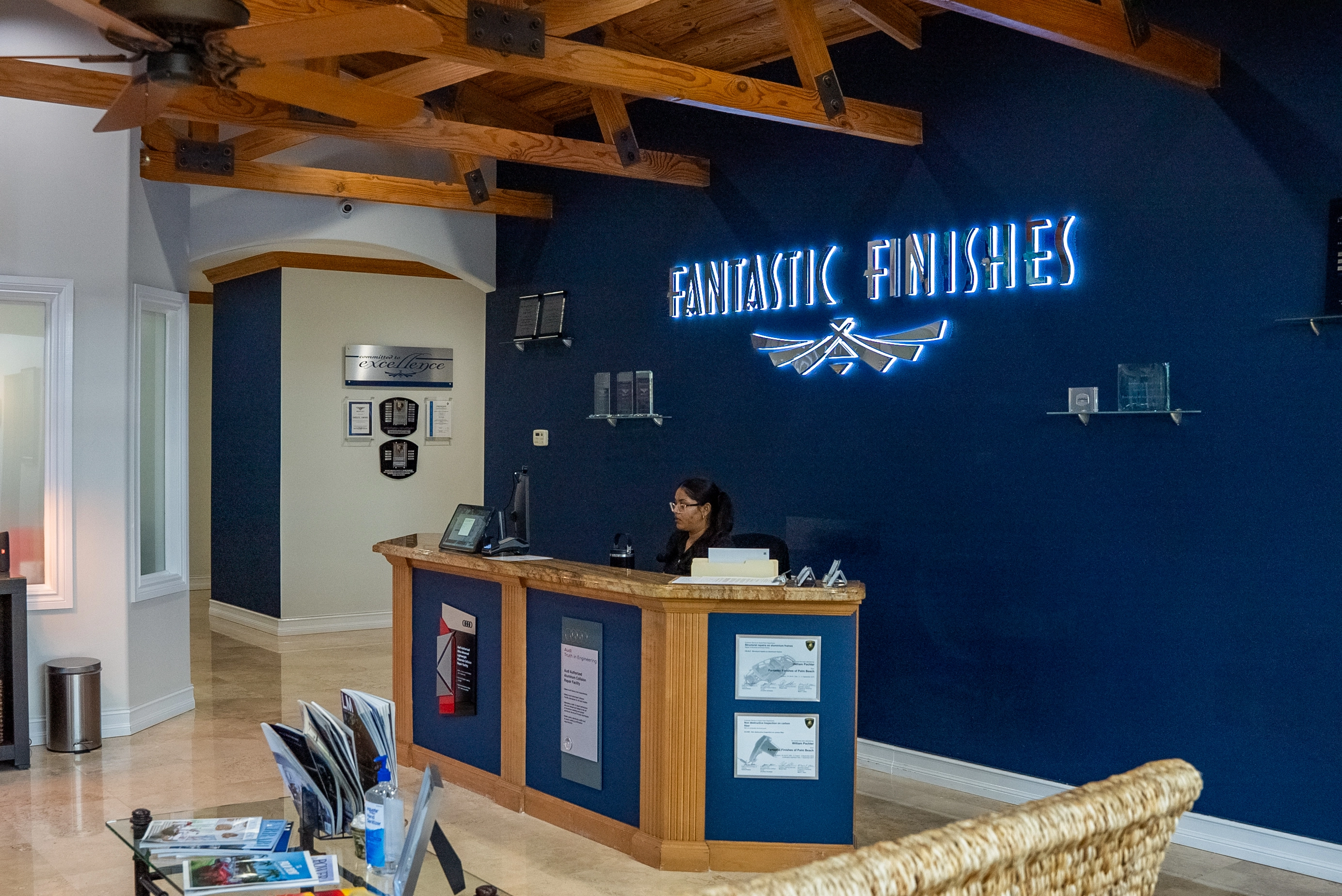 Reception area with a wooden counter where a woman with glasses is working on a computer, in front of a dark blue wall featuring illuminated text reading 'FANTASTIC FINISHES' and a logo, with wooden ceiling beams and a ceiling fan above.