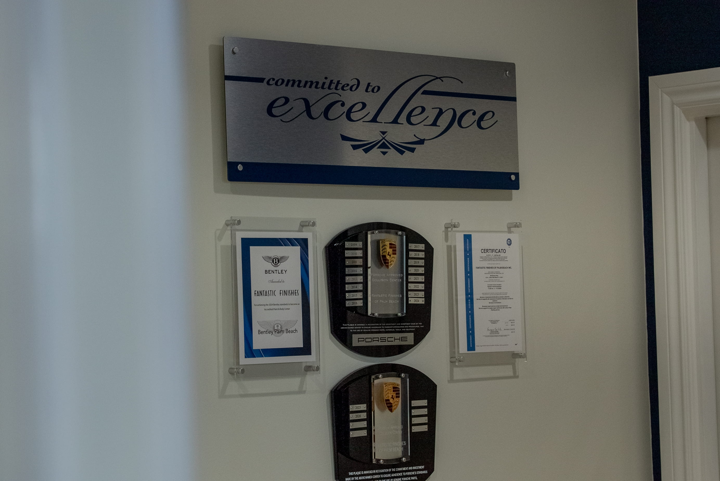 A wall display featuring a metal sign with 'committed to excellence' above two Porsche plaques and two framed certificates, one from Bentley, recognizing Fantastic Finishes of Palm Beach.