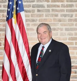 Smiling man in a dark suit with a festive tie stands beside a U.S. flag against a brick wall.