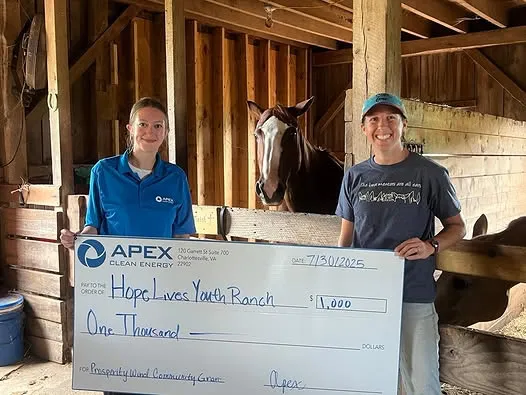 Two people standing in a wooden stable holding a large check from Apex Clean Energy for $1,000 made out to Hope Lives Youth Ranch, with a brown horse visible behind them.