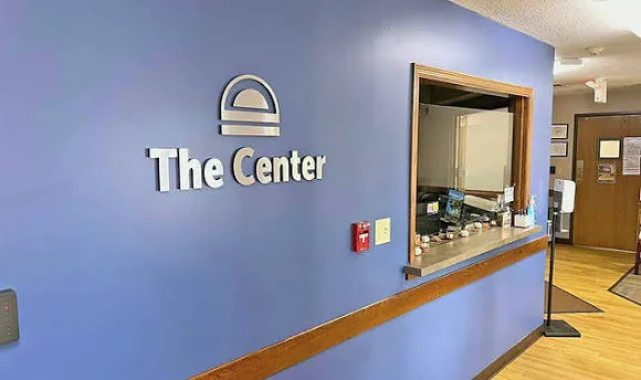 Interior view of a reception area with blue walls featuring a logo and text 'The Center' on the left, a window counter with items on the shelf, a fire alarm, and wooden trimmed doors in the background.