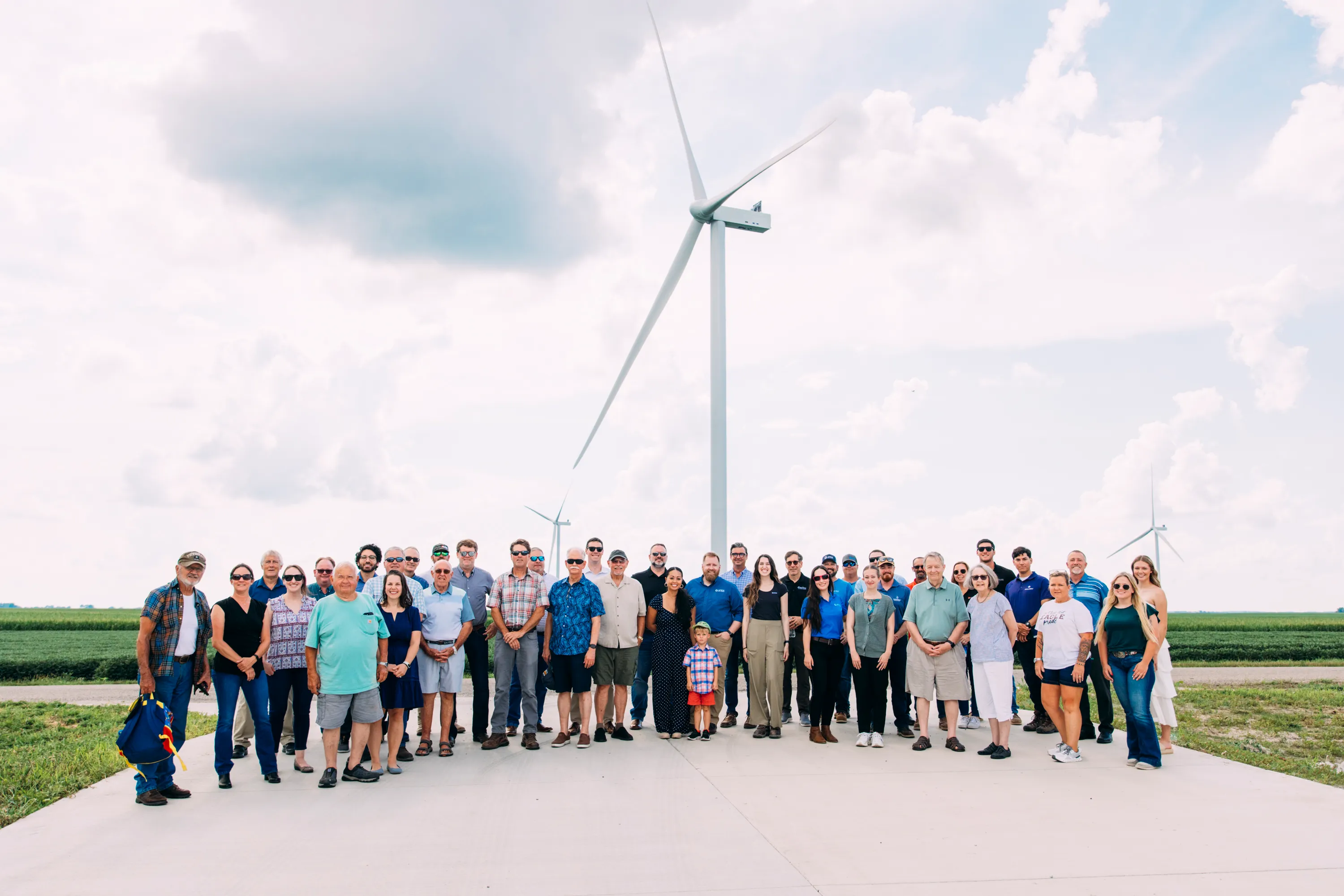A large group of people standing on a concrete surface outdoors with three tall wind turbines in the background under a partly cloudy sky, surrounded by green fields.