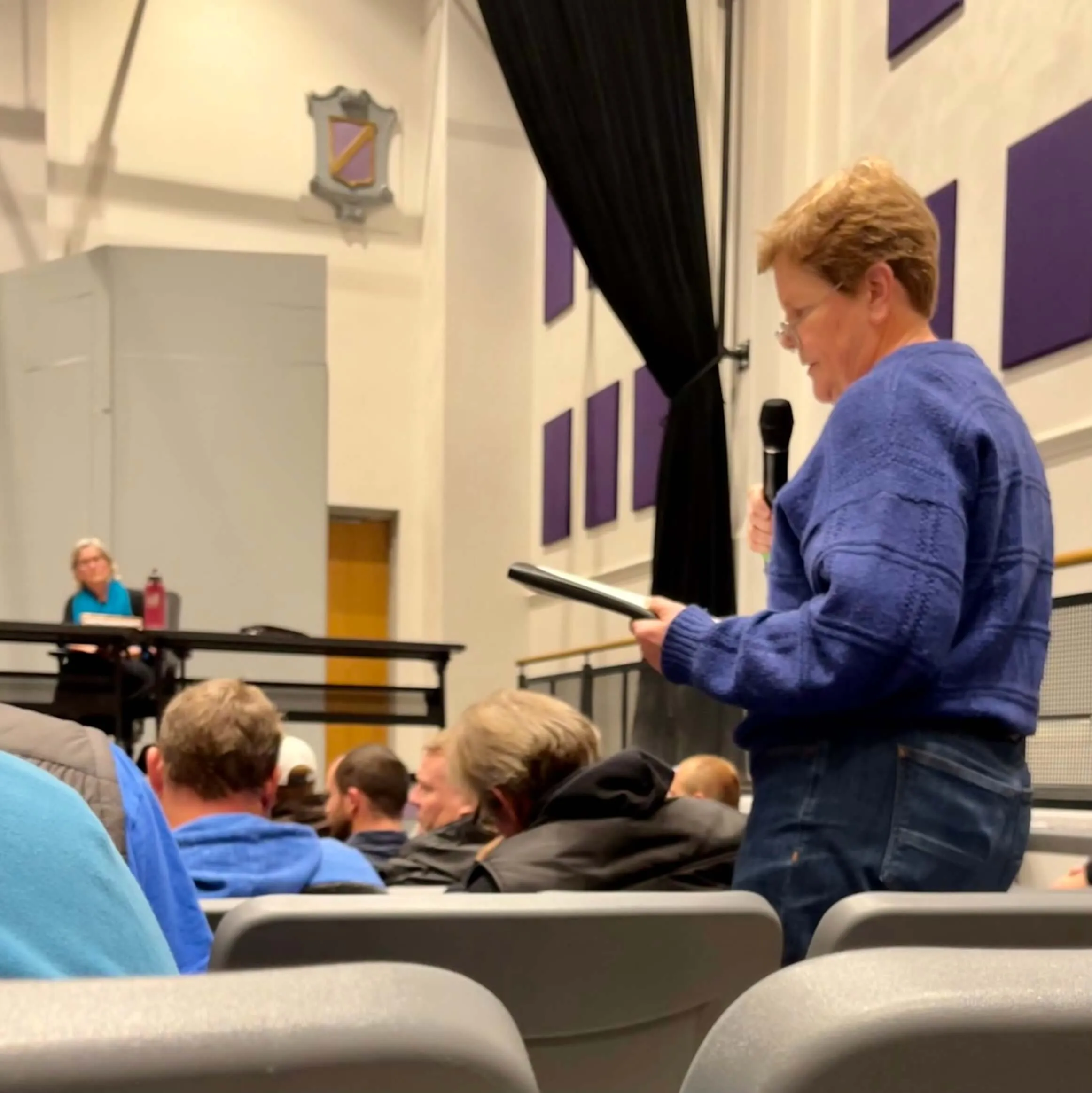 A woman in a blue sweater stands in an auditorium holding a microphone and reading from a tablet, while seated audience members face forward and a woman at a table listens in the background.