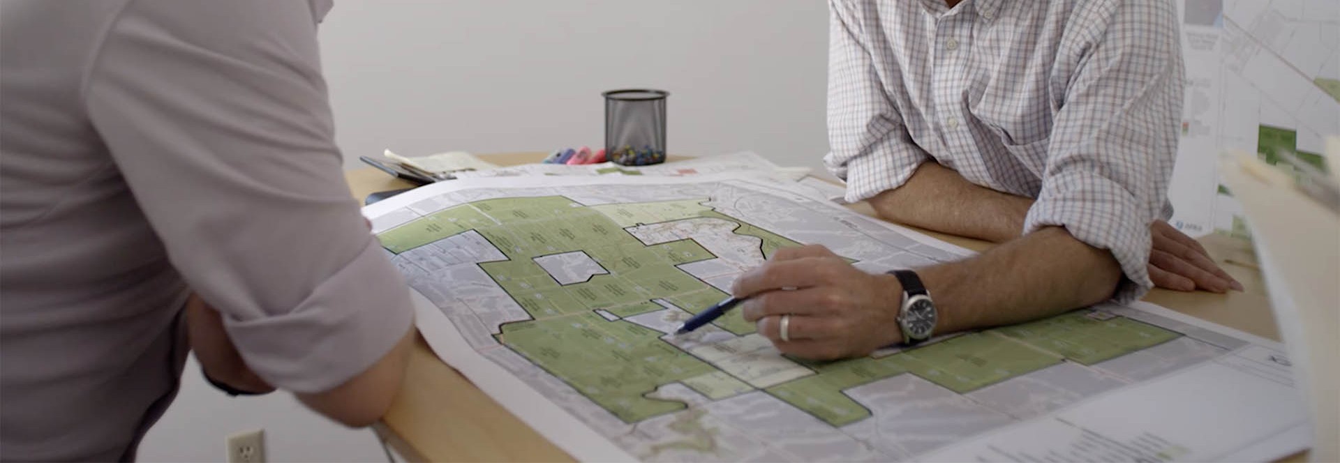 Two men in rolled-up shirts sit at a table examining a large map or project plan with sections highlighted in green; one man points with a pen.
