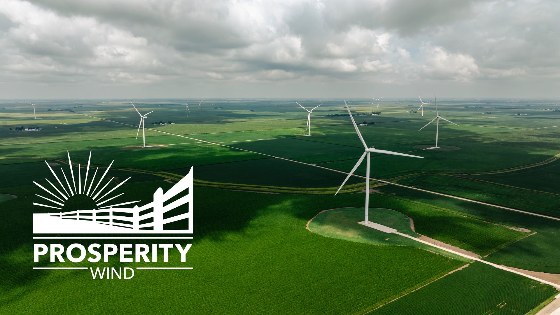Aerial view of a wind farm with multiple turbines over green fields under a cloudy sky.