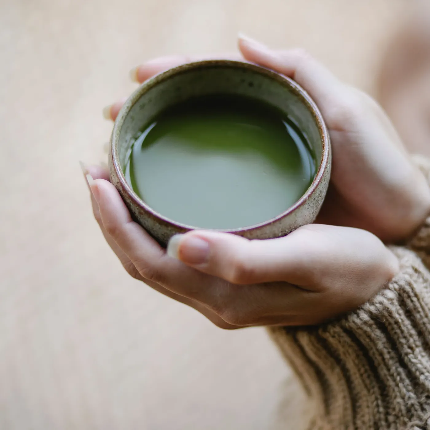 Hands in a knit sweater cradling a stoneware cup of green matcha