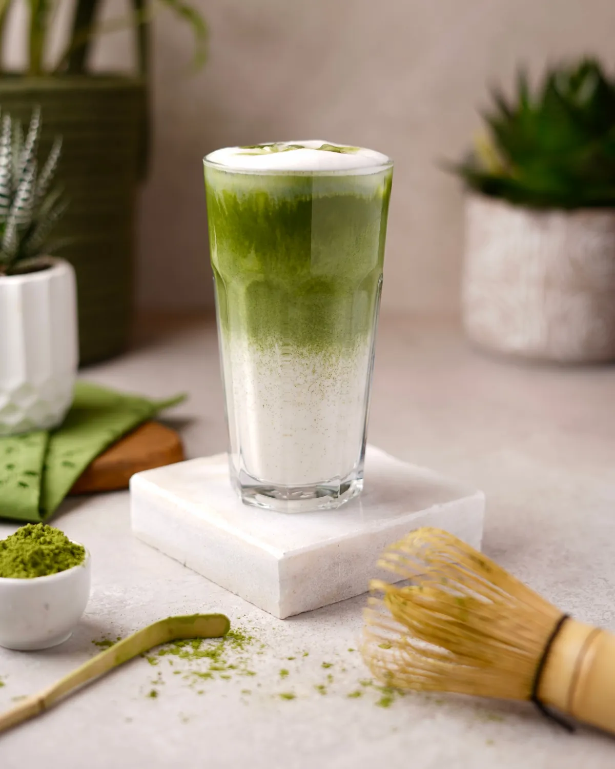 Layered iced matcha latte in a tall glass on a marble block, with a bowl of matcha powder and a bamboo whisk