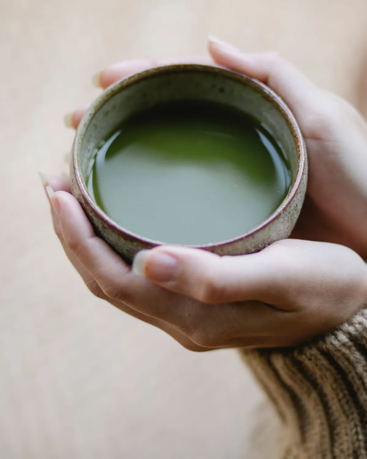 Hands in a knit sweater cradling a stoneware cup of green matcha