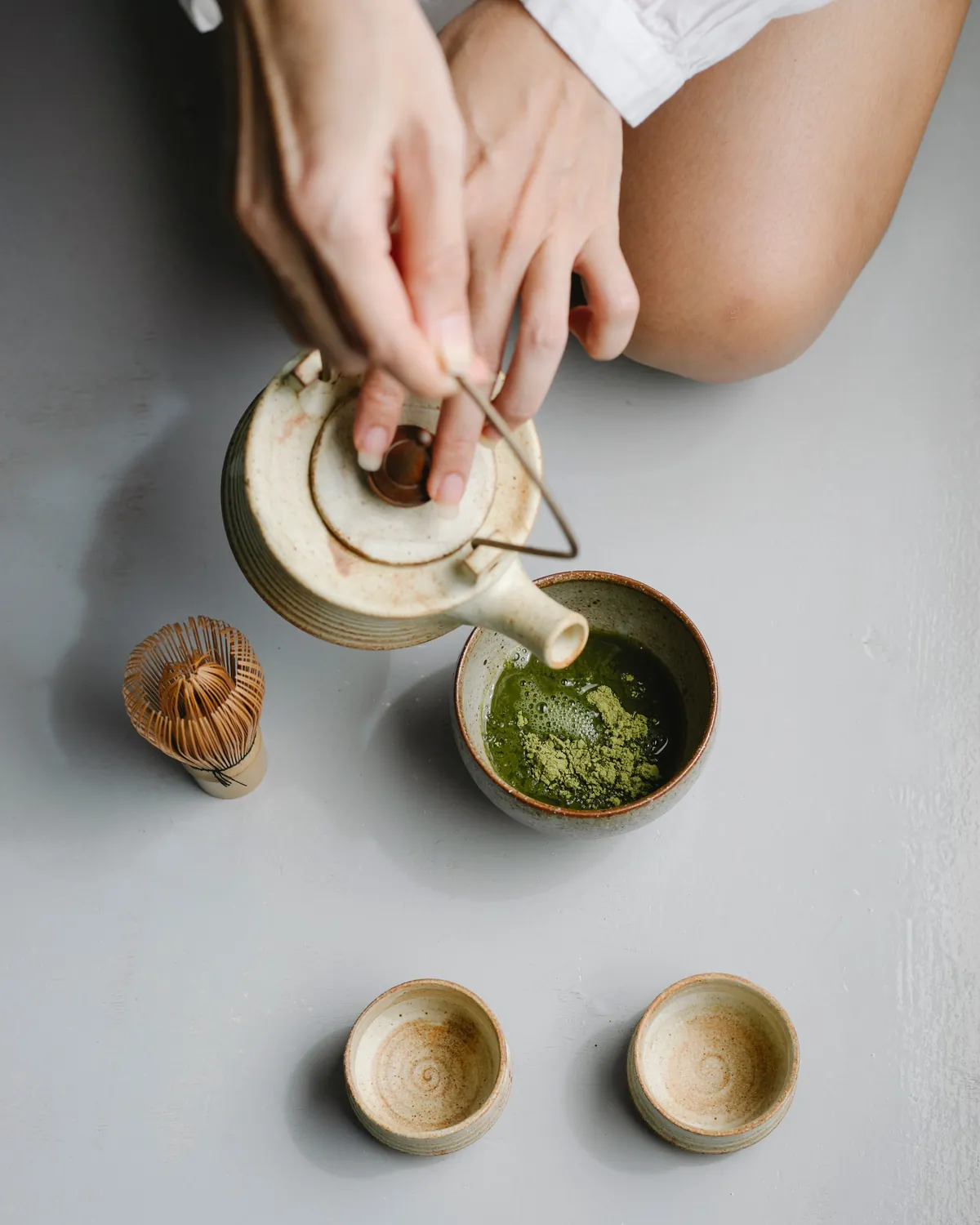 Hands pouring hot water from a stoneware teapot into a bowl of matcha powder