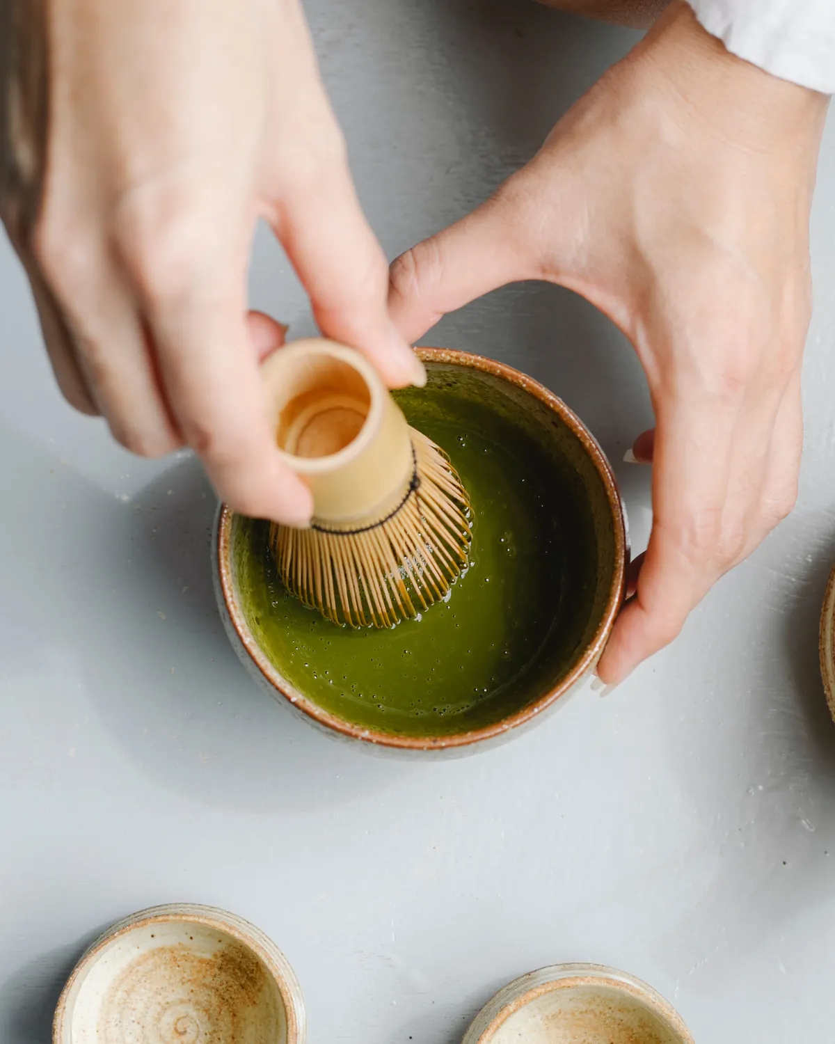 Top-down view of hands whisking bright green matcha in a stoneware bowl