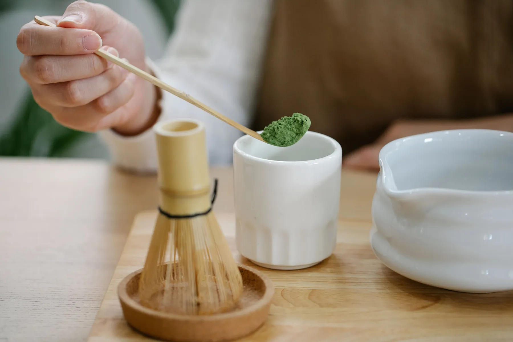 Bamboo scoop of bright green matcha held over a white cup beside a bamboo whisk on a wooden tray