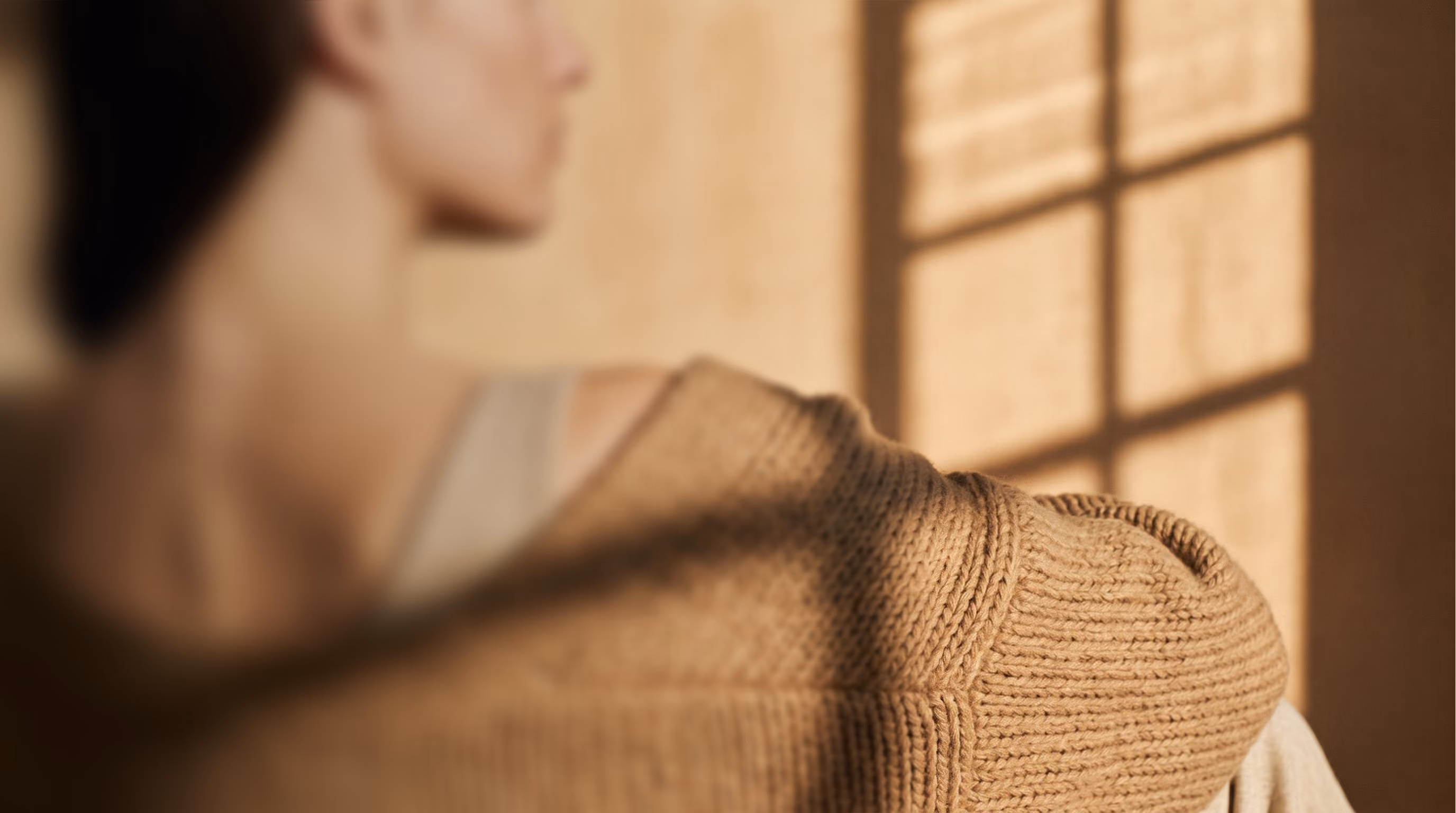 Une femme en pull beige, de dos, dans la lumière d'une fenêtre