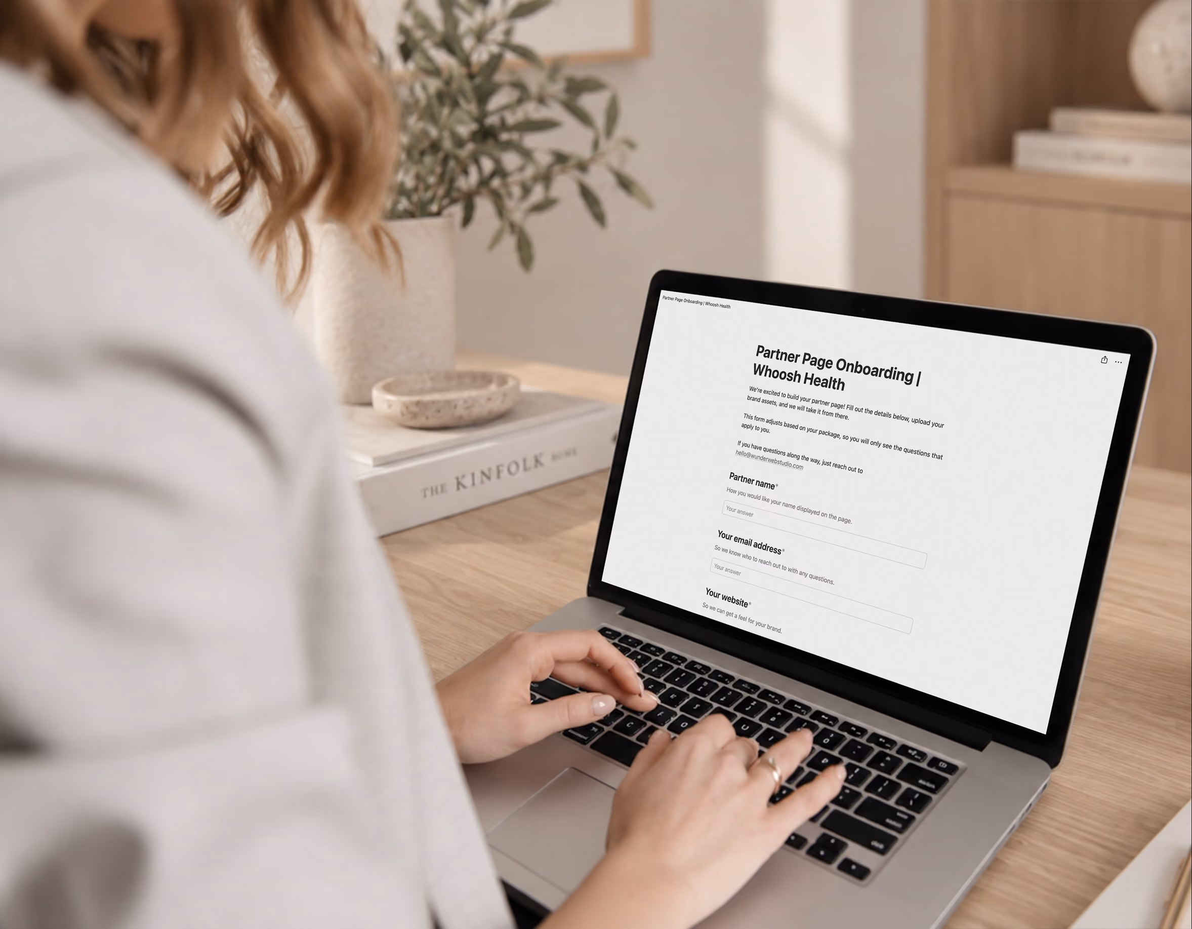 Person typing on a laptop displaying a 'Partner Page Onboarding | Whoosh Health' form with fields for partner name, email address, and website on a wooden desk with a plant and book in the background.