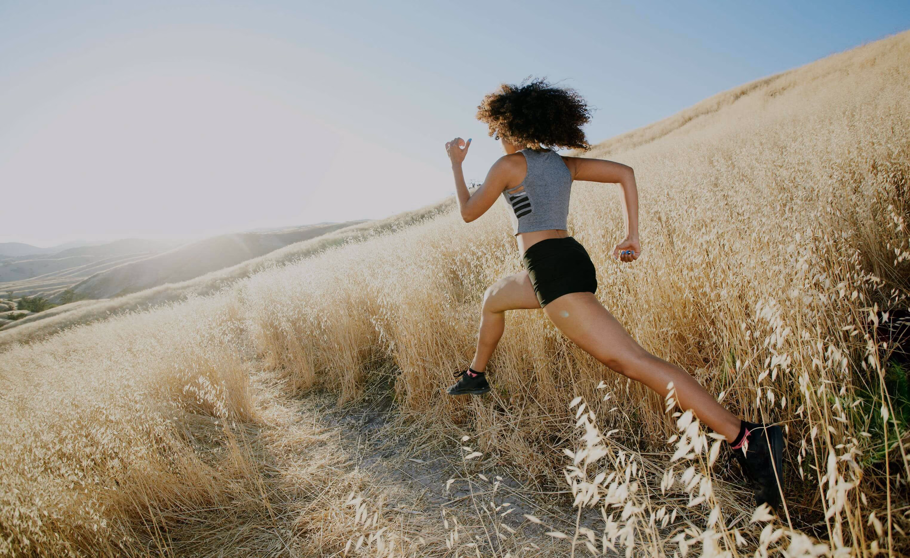A woman with curly hair wearing a grey sleeveless top and black shorts is running uphill on a dirt trail through a dry grassy field under a clear blue sky.