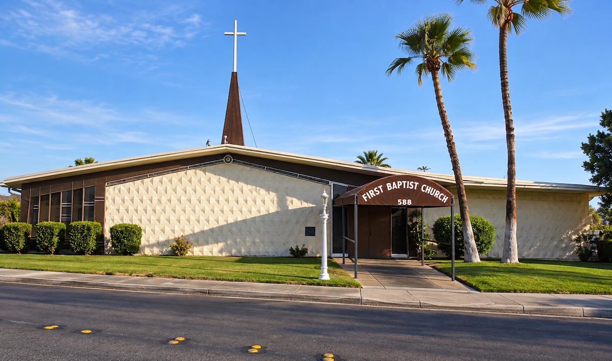 First Baptist Church in Palm Springs: a low building with a textured, patterned concrete facade, a tall spire topped by a cross, and a First Baptist Church sign over the entrance, framed by palm trees