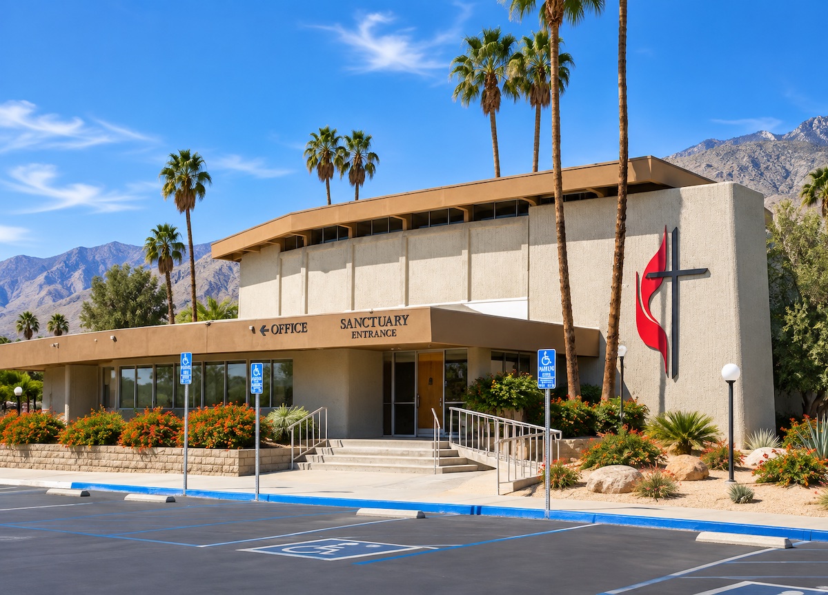 United Methodist Church of Palm Springs: a two-story building with a deep flat roof overhang and a large red cross-and-flame emblem on its wall, beside the sanctuary entrance, with palm trees and mountains behind