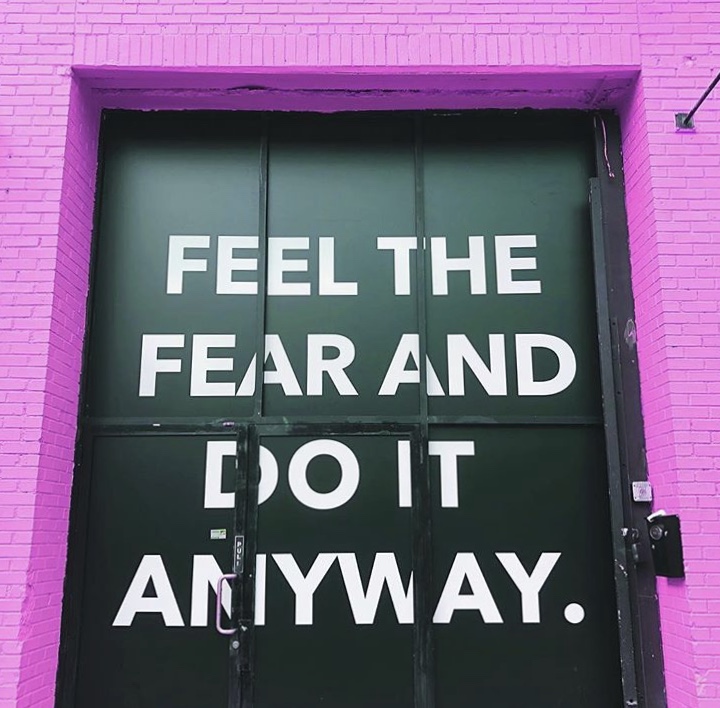 Motivational wall sign reading feel the fear and do it anyway on green doors framed by bright pink brick wall.|