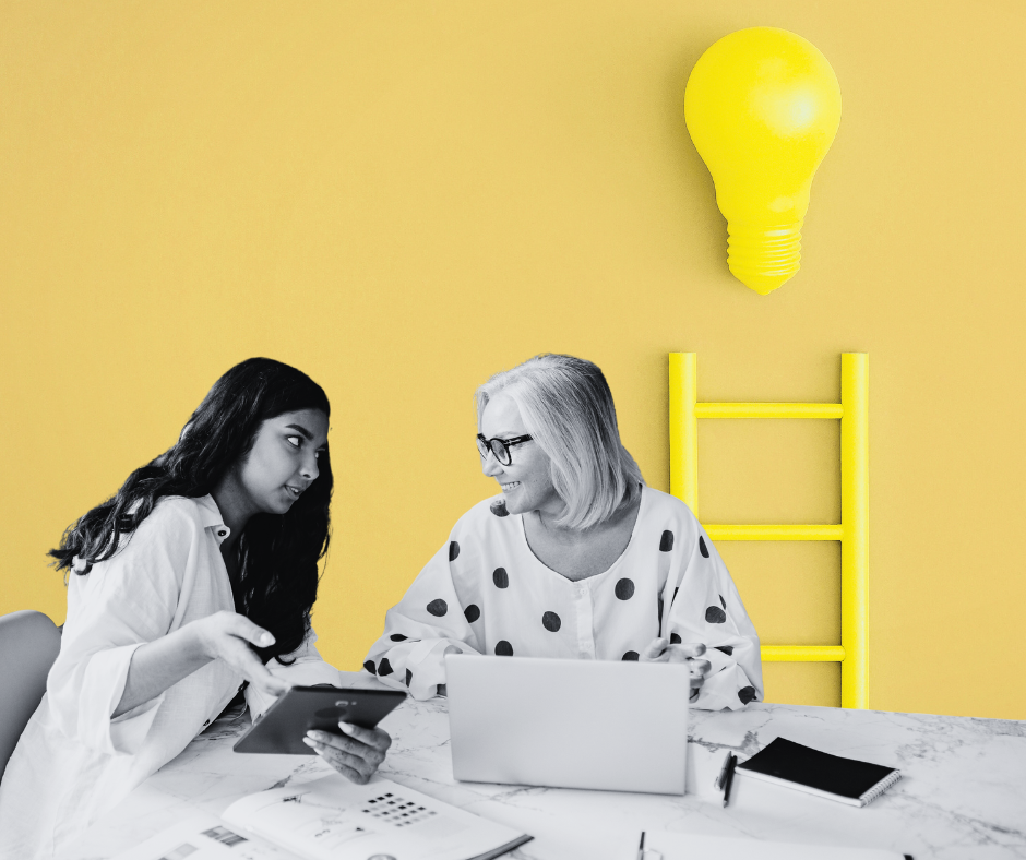 Two women sitting at a desk working, with a ladder and a target in the background.