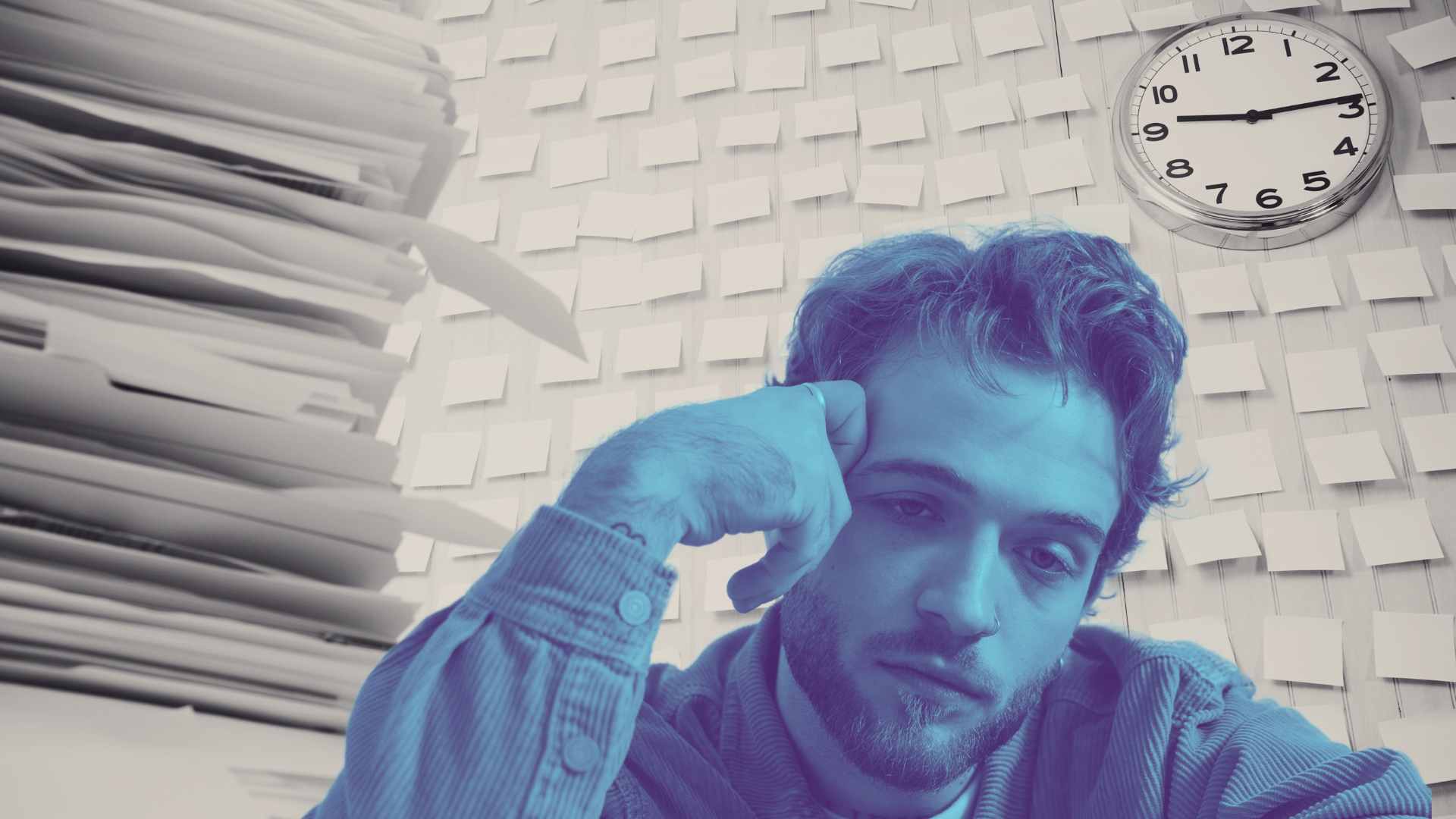 Man sitting at desk with stacks of paperwork, sticky notes on wall, and clock overhead, representing overwhelm, deadlines, and work stress.